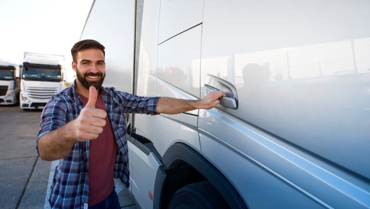 Professional middle aged bearded trucker standing by his semi truck and holding thumbs up.